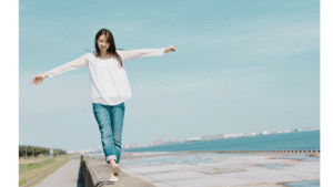A young woman balances with her arms outstretched while walking along a seaside ledge on a sunny day.