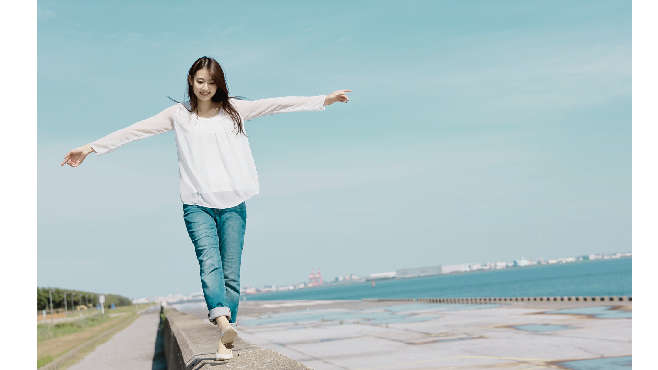 A young woman balances with her arms outstretched while walking along a seaside ledge on a sunny day.