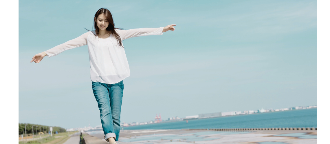 A young woman balances with her arms outstretched while walking along a seaside ledge on a sunny day.